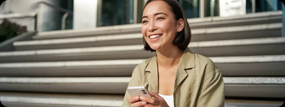 Sara, smiling outdoors with her phone in natural light